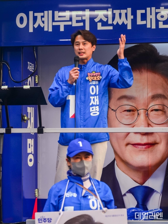 더불어민주당 황대호 청년대변인이 거리 유세 현장에서 연단에 올라 발언하고 있다. ‘이제부터 진짜 대한민국’이라는 슬로건이 적힌 유세 차량 앞에서 마이크를 들고 손짓하며 청중에게 메시지를 전하고 있다.