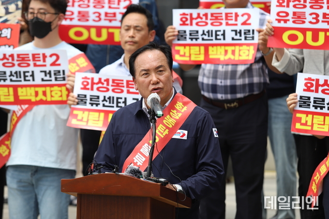이권재 오산시장이 6월 19일 화성시 동탄호수공원에서 열린 초대형 물류센터 반대 기자회견에서 발언하고 있다. 뒤편에는 '화성동탄2 물류센터 건립 백지화'를 요구하는 시민들이 피켓을 들고 함께 반대 의사를 표시하고 있다.