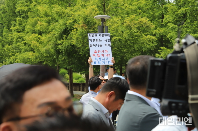 화성시 동탄호수공원에서 19일 열린 초대형 물류센터 반대 집회 현장에서, 물류센터 추진 업체 관계자가 '적법한 사업 진행, 지연되는 사업 늘어나는 피해. 오산시가 책임지나? 화성시가 책임지나?'라는 문구가 적힌 팻말을 들어 보이며 입장을 밝히고 있다.