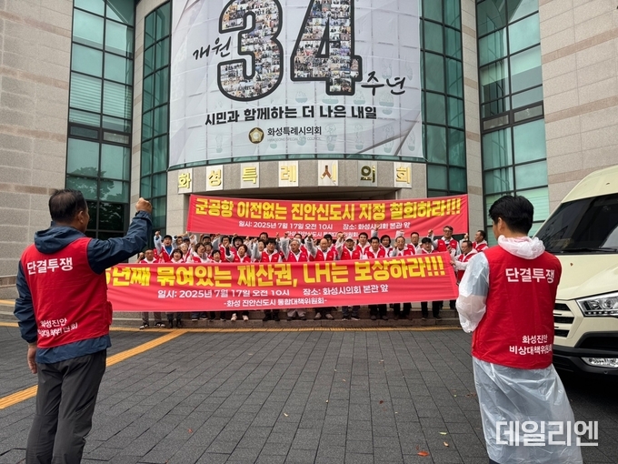 화성시의회 본관 앞에서 17일 열린 ‘화성진안지구 통합대책위원회’ 집회에서 주민들이 “군공항 이전 없는 진안신도시 지정 철회”와 “5년째 묶여 있는 재산권 보상”을 촉구하며 구호를 외치고 있다. 참석자들은 ‘단결투쟁’ 조끼를 입고 대형 현수막을 펼치며 개발 정책의 전면 재검토를 요구했다.